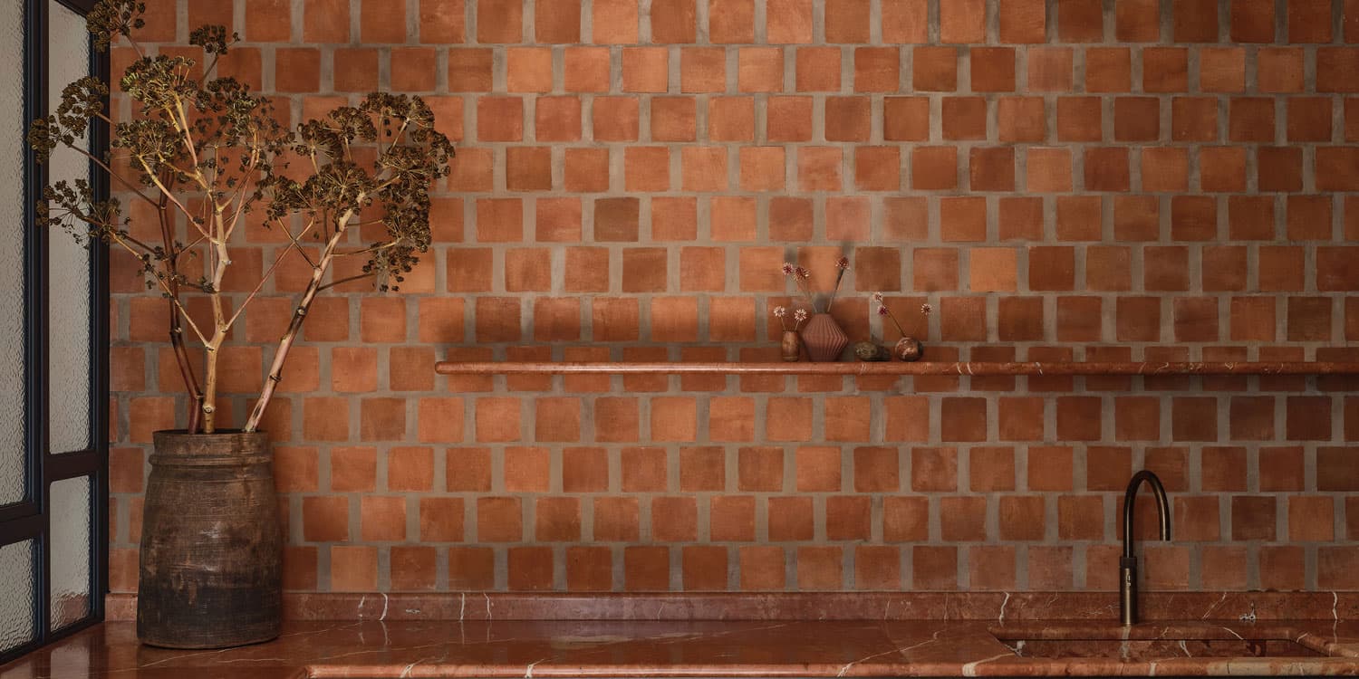 A bathroom with warm, terracotta square tiles on the walls, a matching countertop, a dark sink faucet, dried branches in a large rustic vase, and small pottery vases on a shelf.