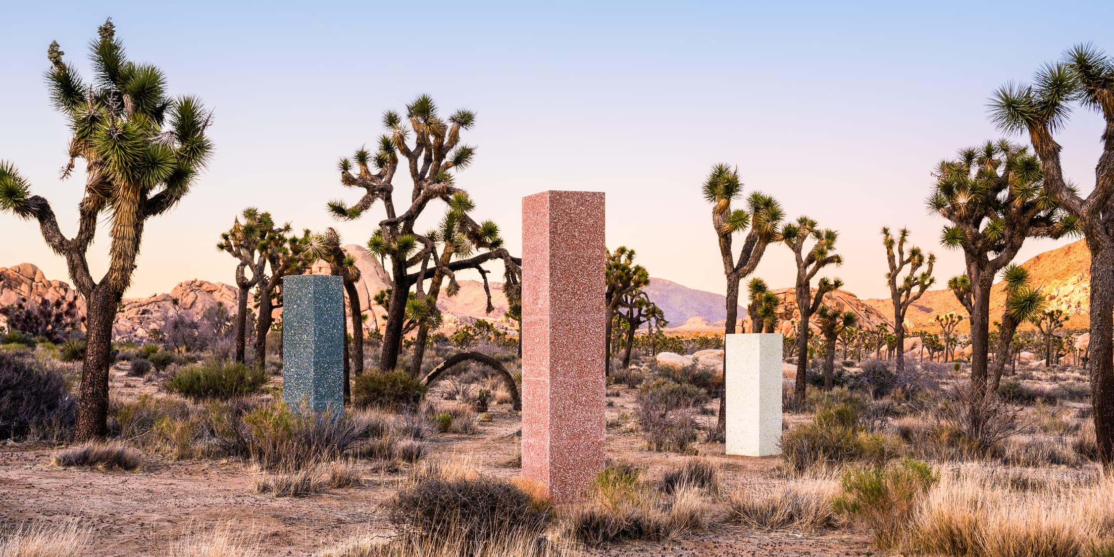 Three tall rectangular monoliths—red, blue, and white—stand in a dry desert landscape with Joshua trees, under a clear sky with mountains in the distance.