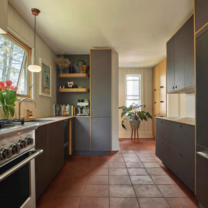 A modern galley kitchen with dark cabinets, terracotta tile floor, wood accents, open shelving with books and decor, a potted plant by a sunlit window, and a stove on the left. Tulips sit on the counter under a window.
