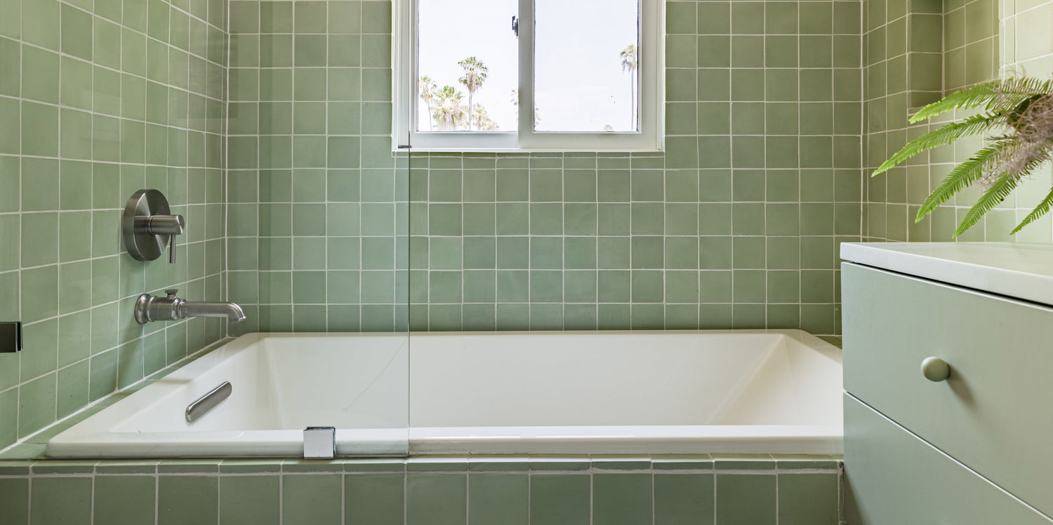 Modern bathroom with green square tiles, a white rectangular bathtub, a wall-mounted faucet, a window above the tub, and part of a green cabinet with a potted plant visible on the right.
