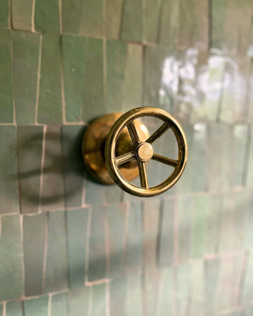 A brass wheel-shaped shower handle is mounted on a wall of Zia Tile’s Absinthe Trapezoid, a glossy green textured Moroccan tile.