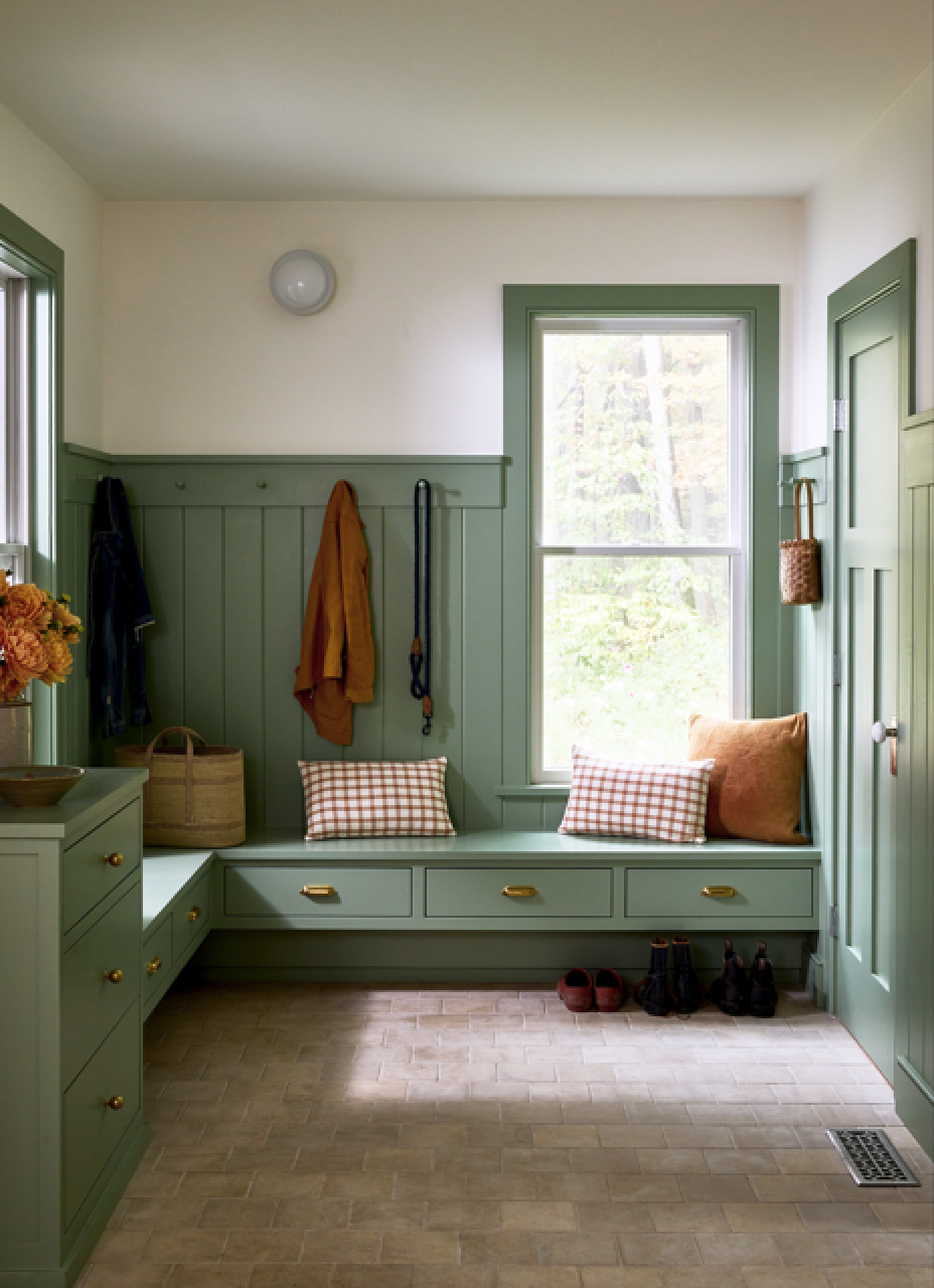 A cozy mudroom with sage green built-in bench and cabinets, checkered and solid pillows, baskets, hooks with dog leashes, and shoes on a tiled floor. Large windows let in natural light.