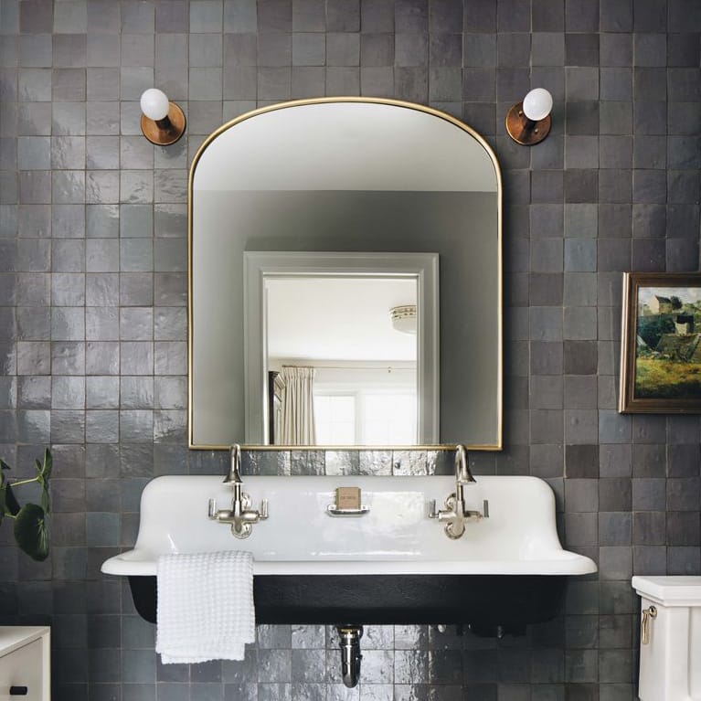 A modern bathroom with glossy Graphite Grey 4x4 tiles by vendor-unknown, a large arched gold-framed mirror, dual wall sconces, a vintage-style sink, white towel, and a framed picture on the wall.