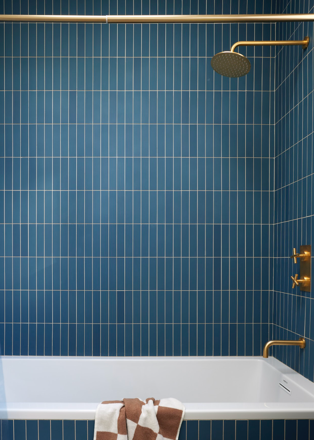 A modern bathroom with a white bathtub, blue vertical tiled walls, gold shower fixtures, and a brown and white striped towel draped over the tub.