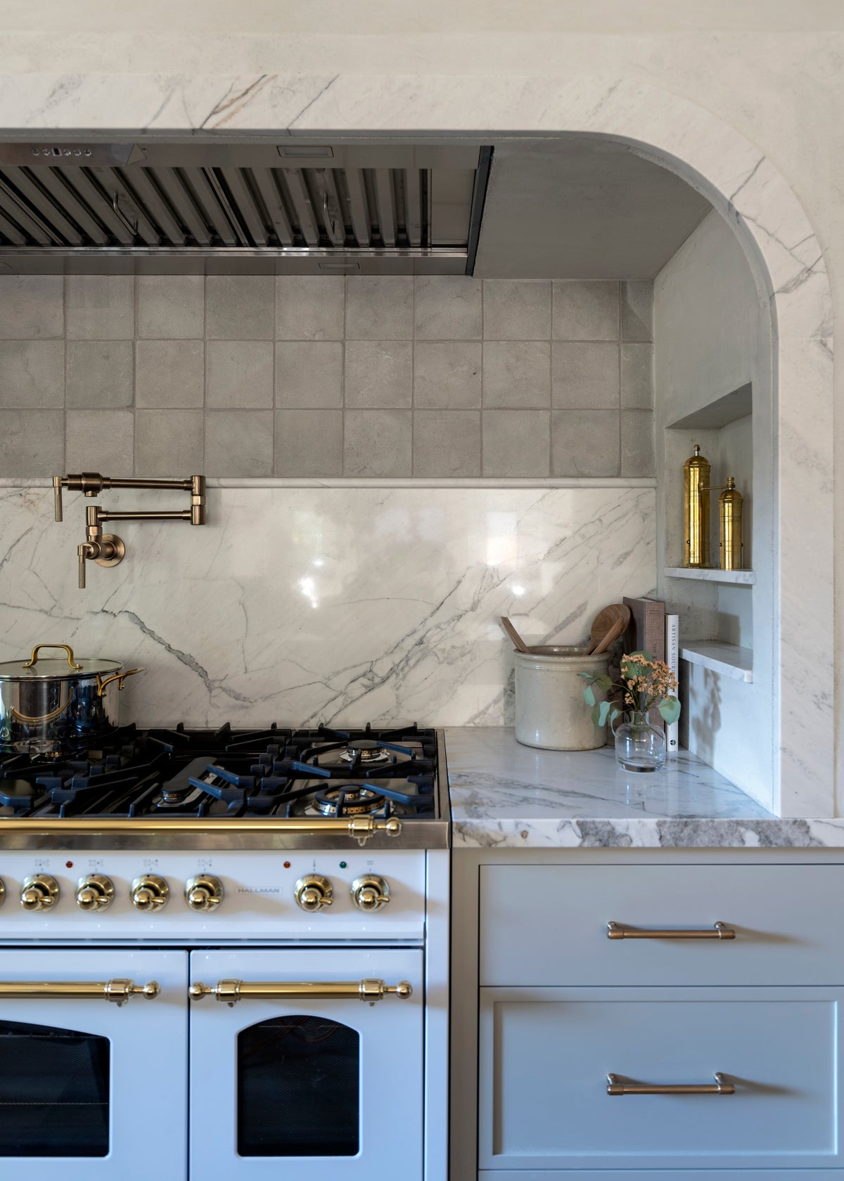 A modern kitchen with a white and gold stove, marble backsplash and countertop, a pot on the stove, a brass pot filler, and built-in shelves holding bottles, a bucket, and a small vase with flowers.