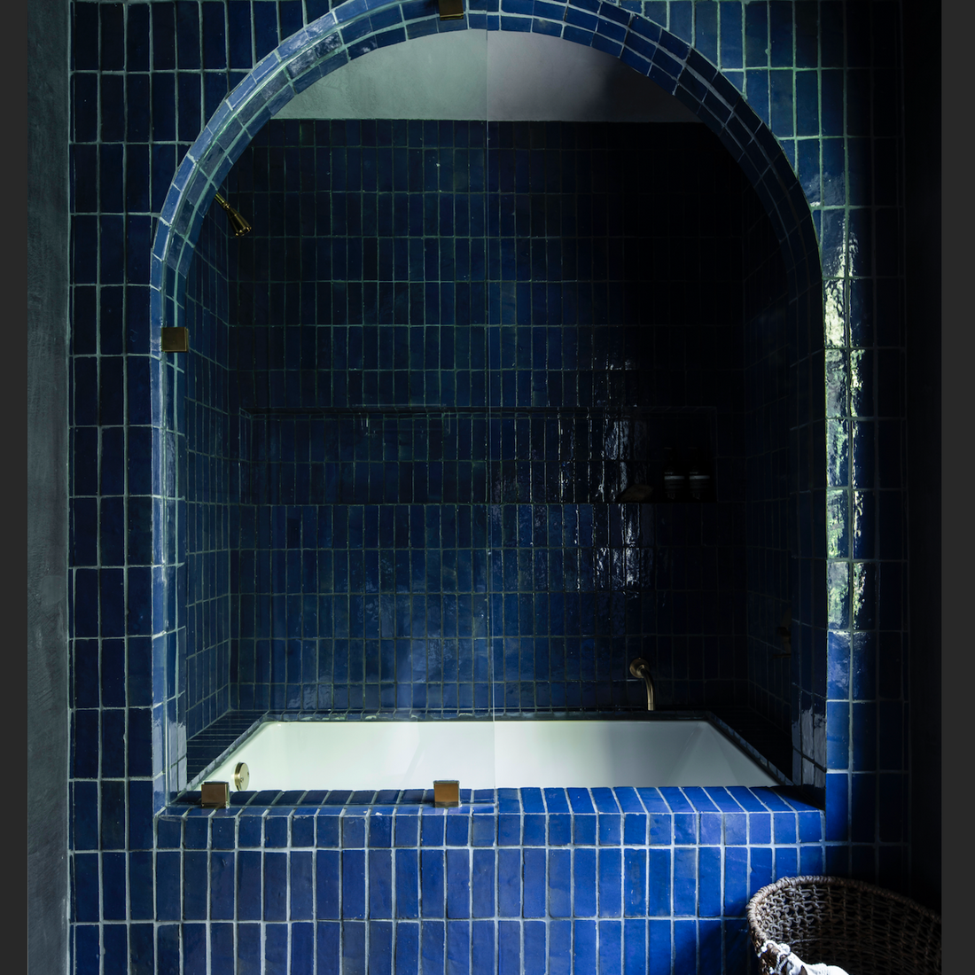 A bathtub is nestled in a nook with Zia Tile's Night Blue 2x6 tiles covering the walls and arch, creating a cozy Moroccan-inspired atmosphere. A wicker basket with a white towel sits nearby under moody lighting.