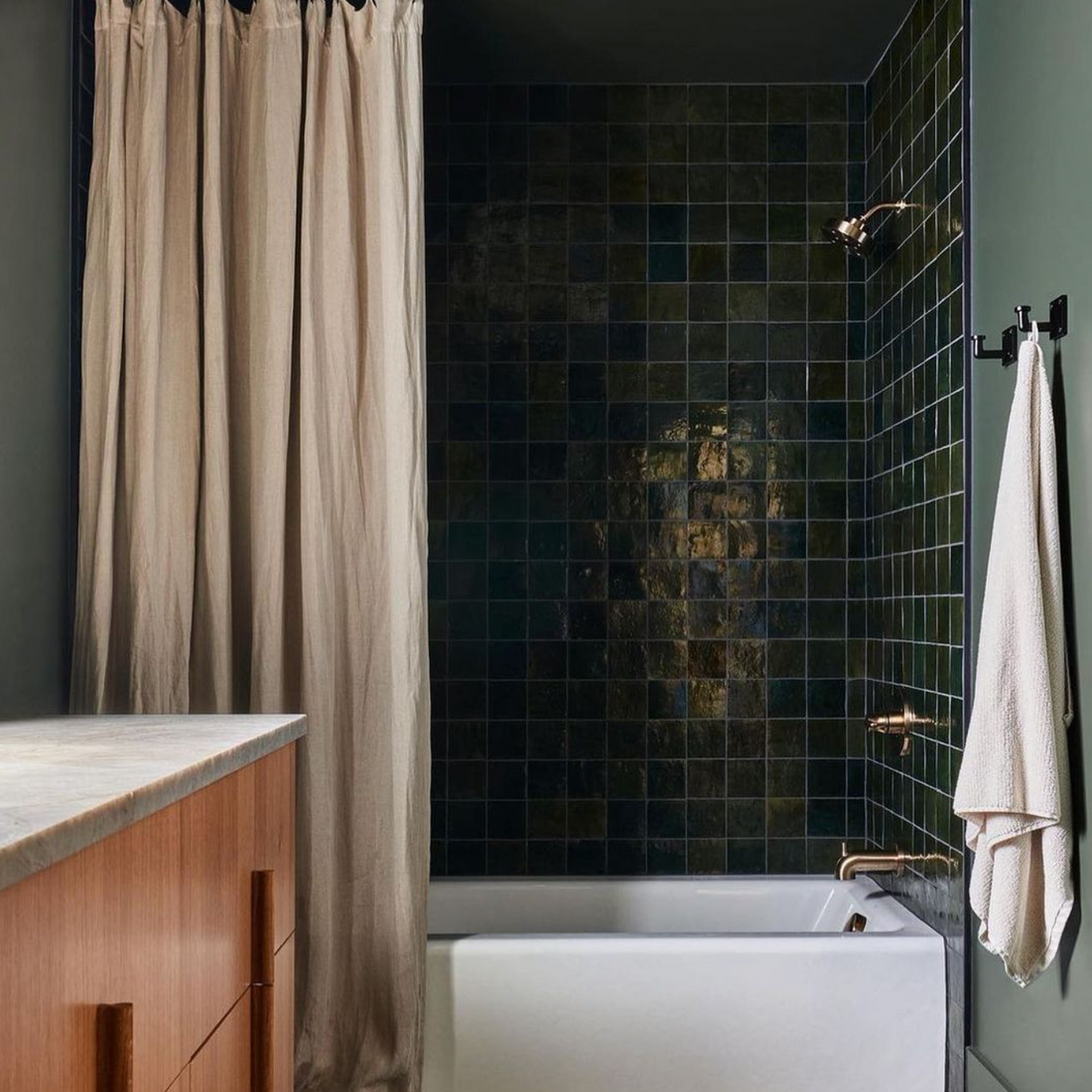 A modern bathroom featuring vendor-unknown Racing Green 4x4 tiles on the walls, a white bathtub, beige shower curtain, brass fixtures, a wooden vanity with marble countertop, and a white towel hanging on a black hook.