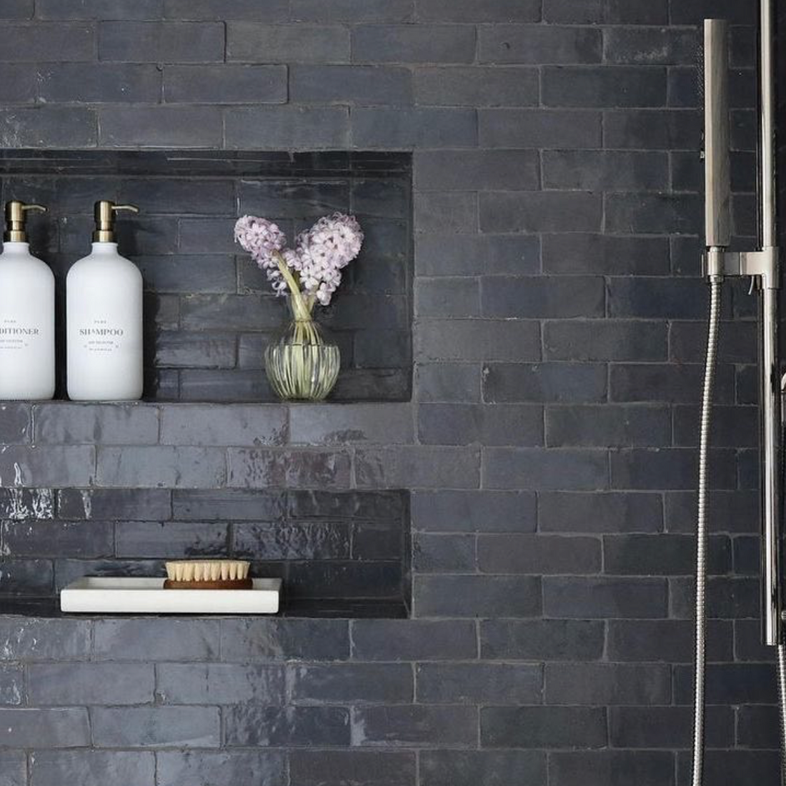 A shower with glossy Slate Grey 2x6 Zia Tile features built-in shelves holding two white pump bottles, a small vase with purple flowers, a bar of soap, and a brush. The Moroccan tile adds elegance, paired with sleek silver fixtures.