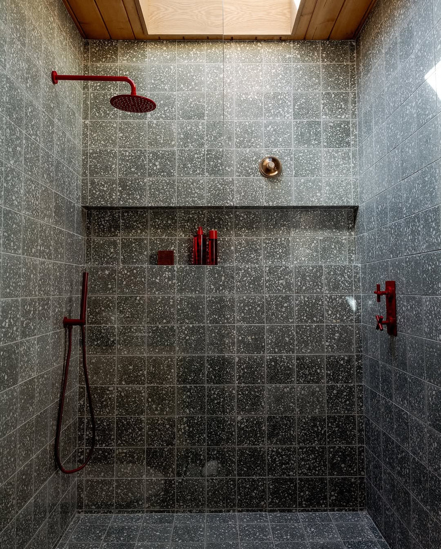 Contemporary shower features Zia Tile’s Smoketree 12x12 gray terrazzo tiles, red fixtures, rain and handheld showers, built-in shelf with bottles, cement flooring, and abundant natural light from the skylight above.
