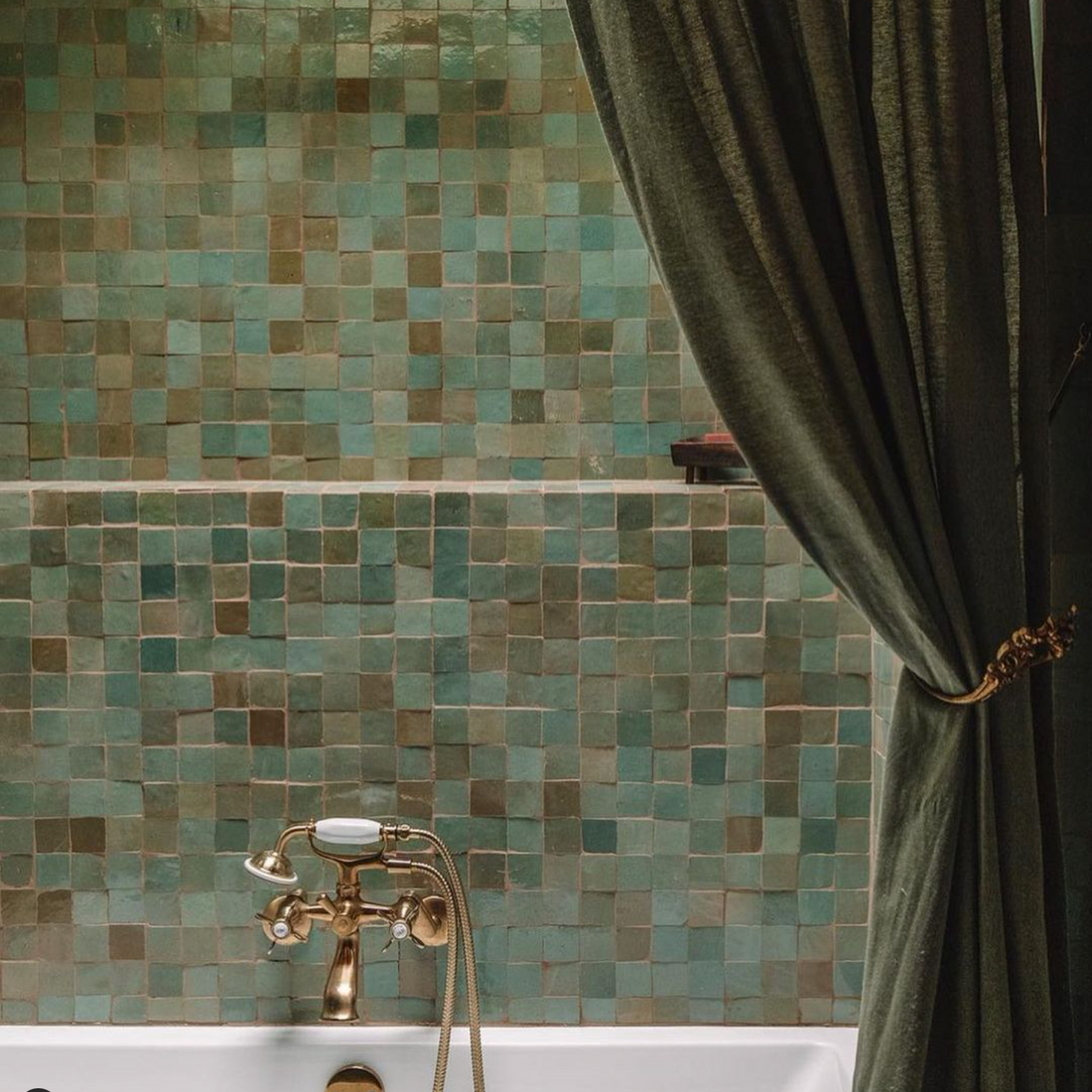 A vintage-style bathtub with a brass faucet sits before a wall clad in Zia Tile’s Tidepool 2x2 tiles, featuring green and teal hues. Dark green curtains to the right enhance the artisanal appeal of this Moroccan-inspired tilework.