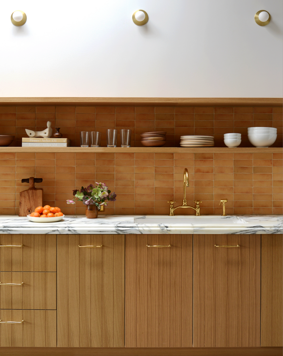 A modern kitchen features wood cabinets, a marble countertop, brass fixtures, and a Zia Tile 2x6 Rectangle + Madera terracotta backsplash. Open shelves hold dishes and glasses; a bowl of oranges sits on the counter.