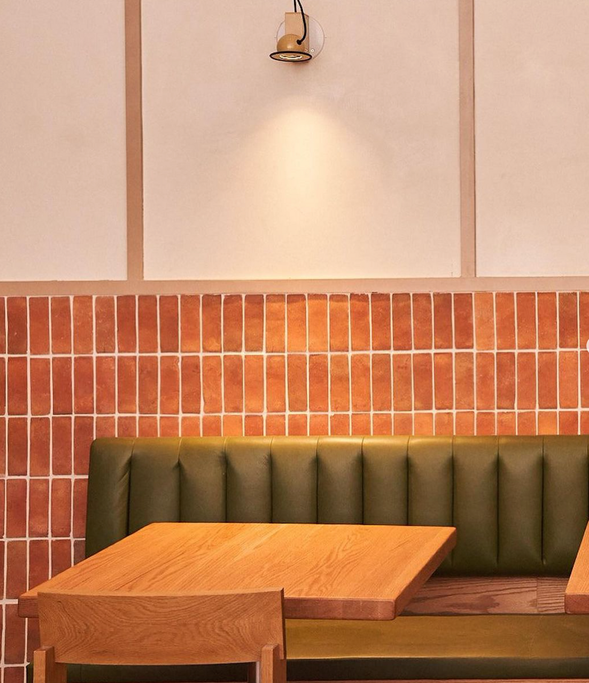 A minimal café interior with a green upholstered bench, wooden tables, and chairs. The wall features a grid of reddish-brown tiles and cream panels, with a single light fixture above the seating area.