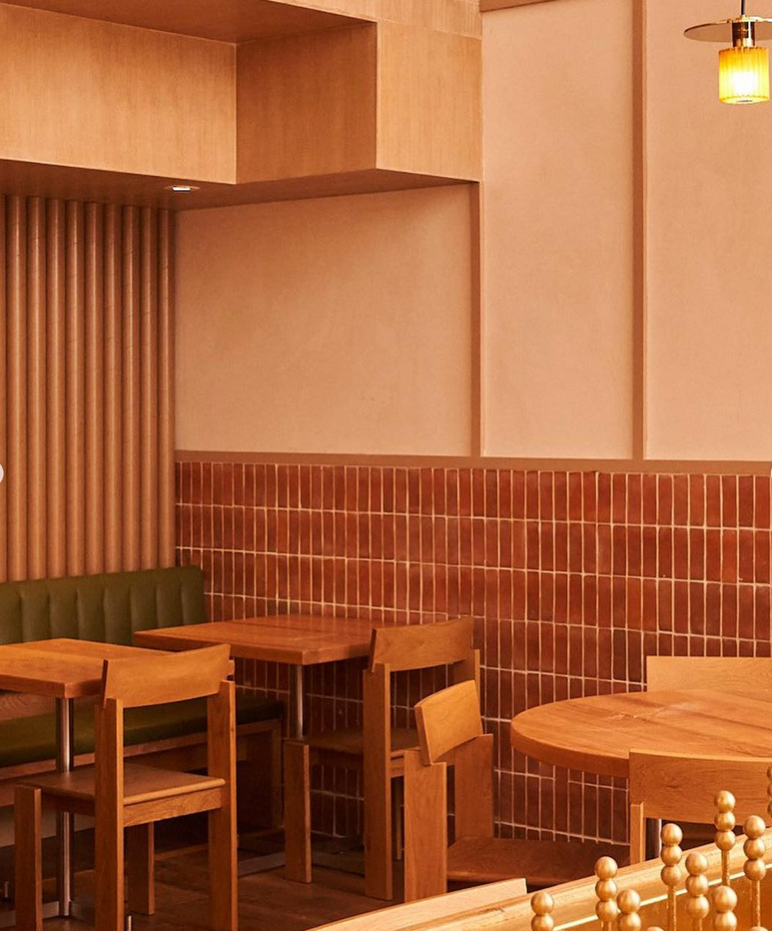 A cozy cafe interior with wooden tables and chairs, green cushioned bench seating, and warm-toned tile and wood panel walls. A small ceiling light adds a soft, inviting glow to the minimalist decor.