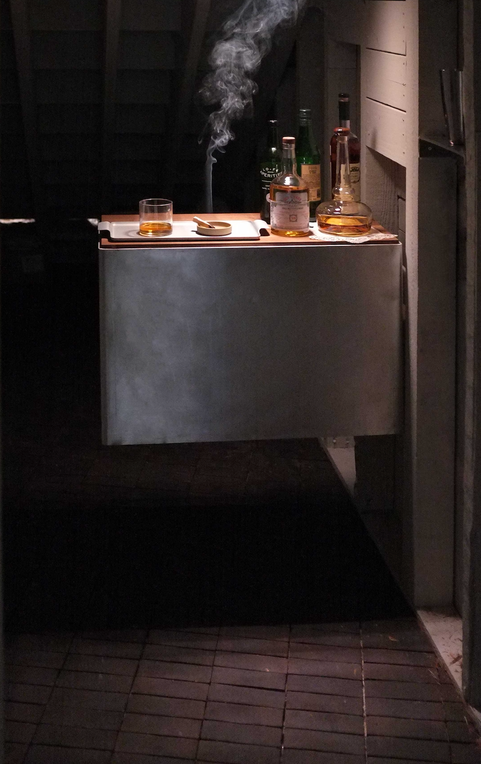 A dimly lit bar scene features Zia Tile’s 2x6 Rectangle + Oscura sample on the counter, with bottles of liquor, a glass of whiskey, a closed book, and a lit cigarette in an ashtray as smoke ascends.