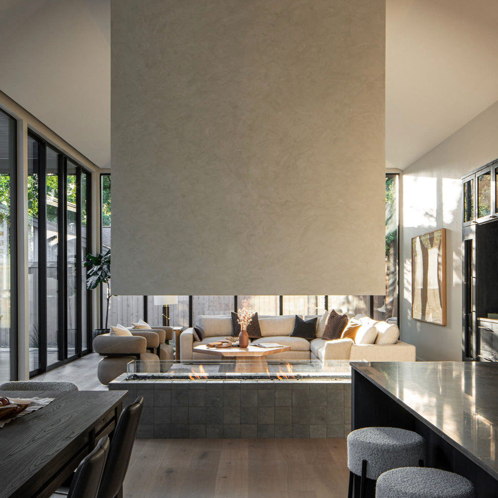 A modern living room with tall windows, a suspended fireplace, a beige sectional sofa, and neutral decor. Sunlight filters in, highlighting minimalist furniture and a cozy, open layout.