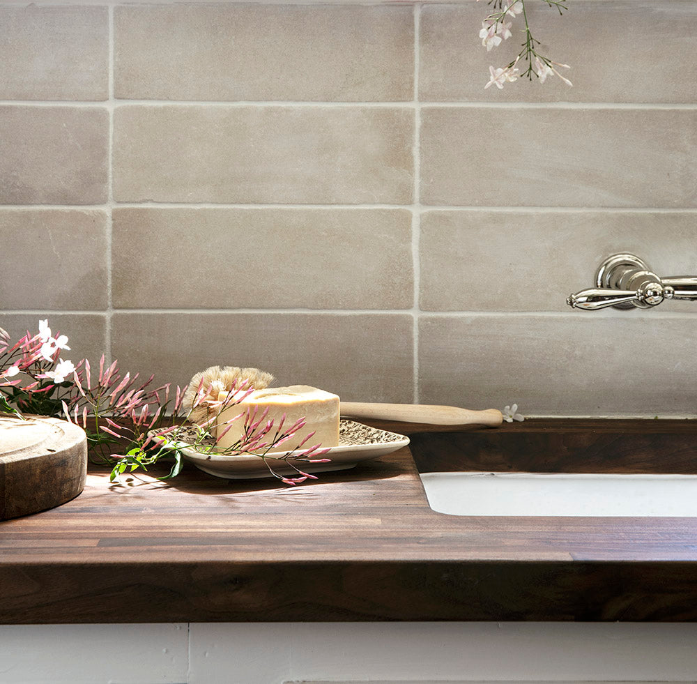A rustic bathroom sink with a wooden countertop, a floral arrangement, a bar of soap on a dish, and a bath brush. Light beige tiles and a silver faucet are visible in the background.