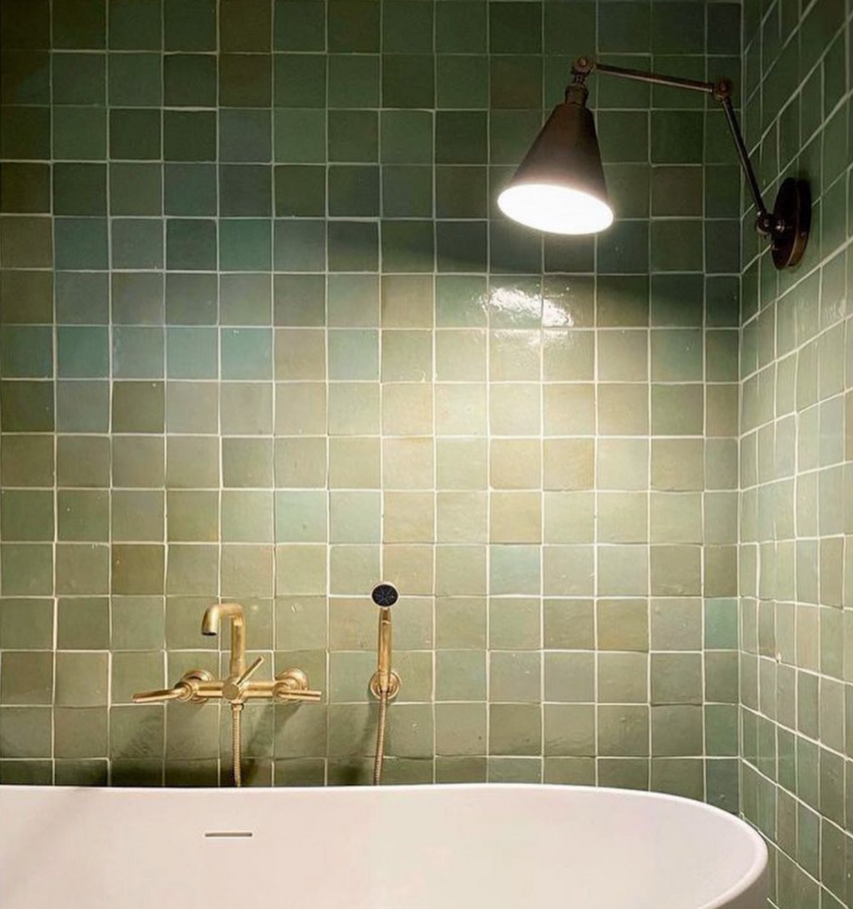 A white bathtub sits against a wall of Tidepool 4x4 tiles by vendor-unknown. Brass fixtures and a black, wall-mounted adjustable sconce with a cone-shaped shade cast warm light onto the green zellige tiles above the tub.