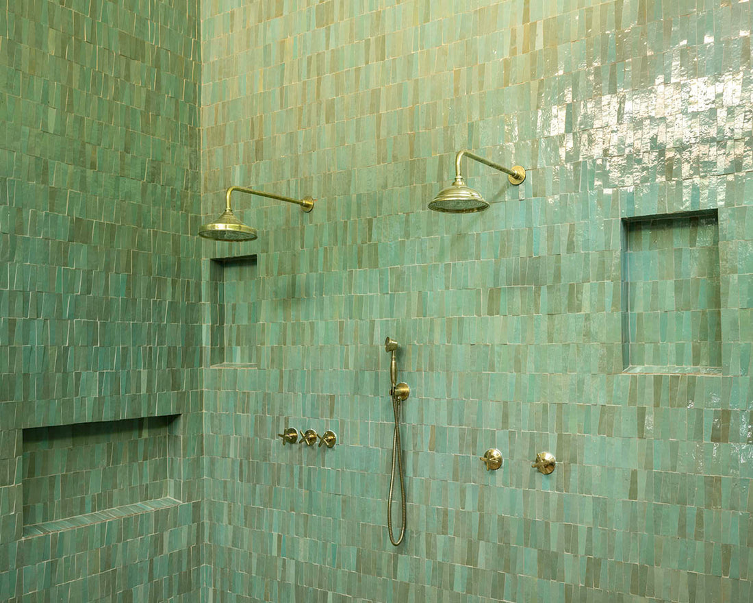 A shower featuring Zia Tile’s Tidepool Trapezoid tiles, two wall-mounted showerheads, a handheld shower, three built-in shelves, and multiple metal knobs for water control.