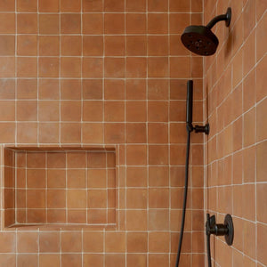 A shower with square, rustic terracotta tiles, black shower fixtures, a handheld showerhead, and a built-in rectangular niche for storage.