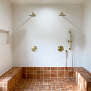 A minimalist shower with two overhead showerheads, brass fixtures, and terracotta-colored tiled bench and floor. The walls are white, and there is a built-in shelf on the left side.
