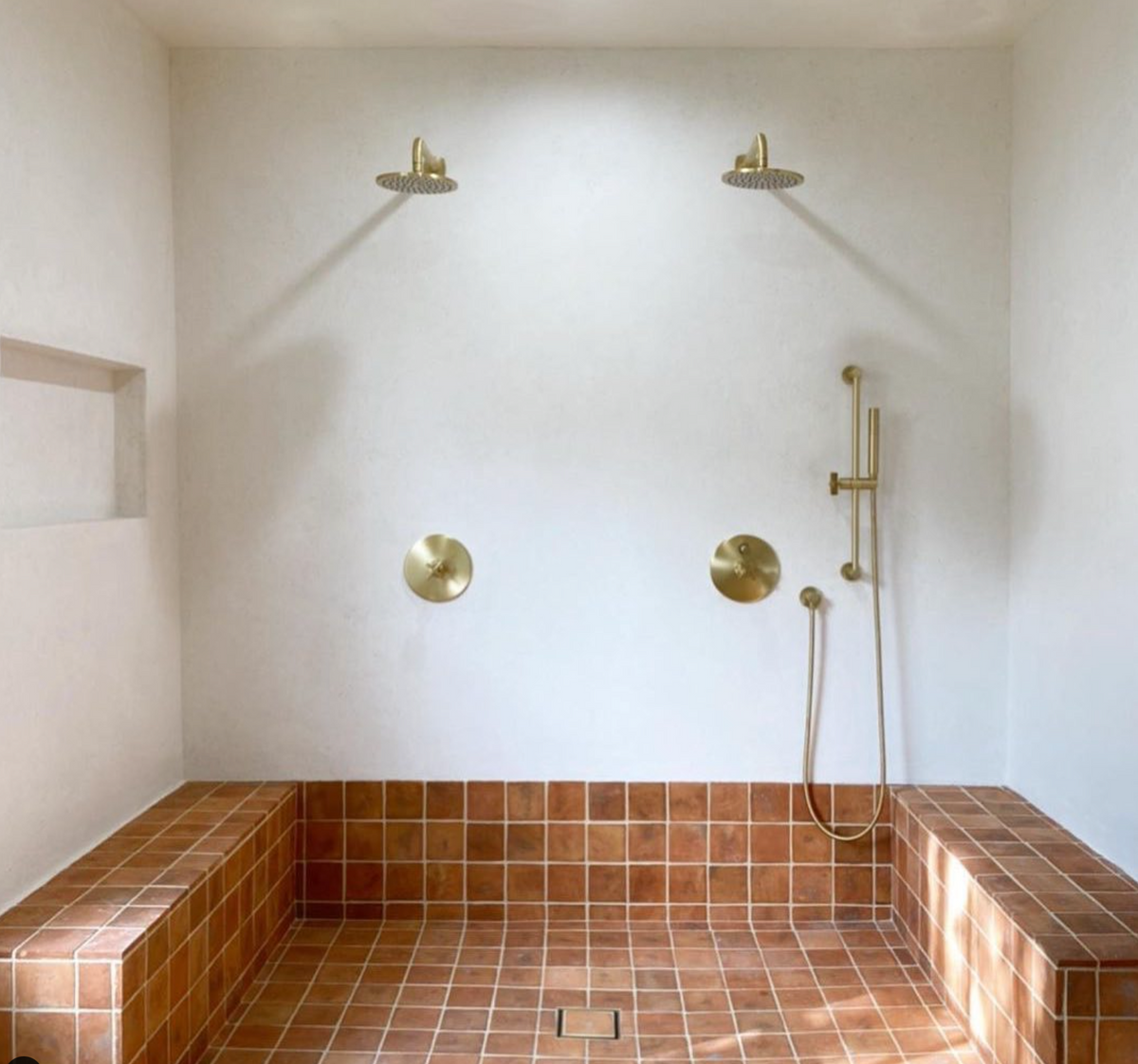 A minimalist shower with two overhead showerheads, brass fixtures, and terracotta-colored tiled bench and floor. The walls are white, and there is a built-in shelf on the left side.