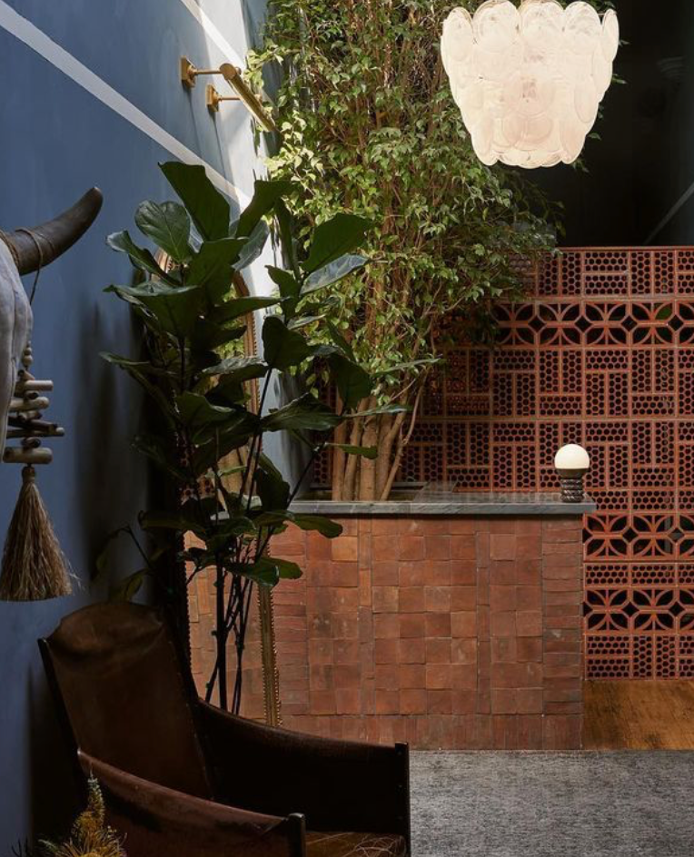 A cozy interior with blue walls, large green plants, a brown leather chair, a tiled counter, a red perforated brick screen, and a white decorative light fixture hanging from the ceiling.