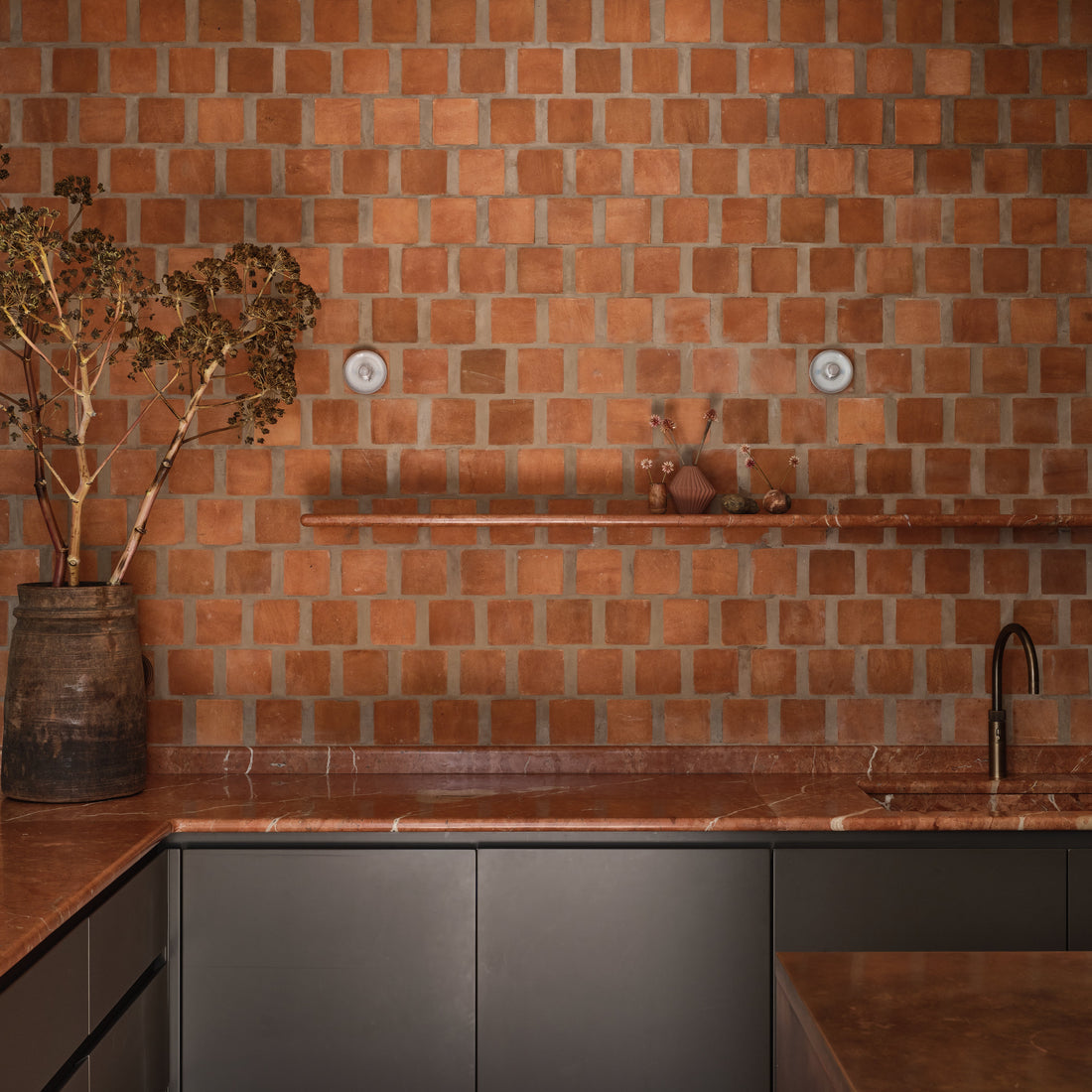 A modern kitchen with reddish-brown square tiles on the wall, a matching countertop, a dark lower cabinet, a minimalist faucet, and a large rustic vase with branches on the left. A narrow shelf holds small decorative items.