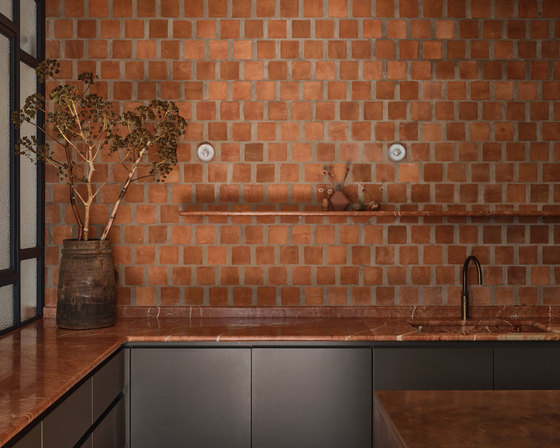 A modern kitchen with reddish-brown square tiles on the wall, a matching countertop, a dark lower cabinet, a minimalist faucet, and a large rustic vase with branches on the left. A narrow shelf holds small decorative items.