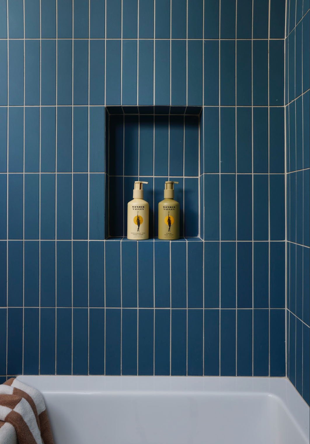 Two yellow soap dispensers are placed in a recessed shelf within a wall of blue vertical tiles above a white bathtub. A brown and white striped towel hangs over the edge of the tub.