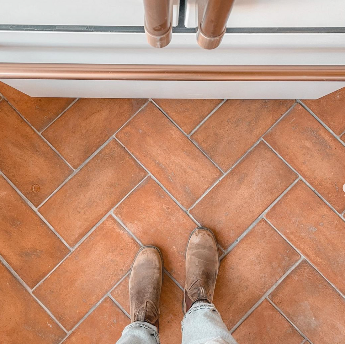 A person wearing light blue jeans and brown boots stands on a terracotta herringbone tile floor in front of a white appliance with copper-colored handles.
