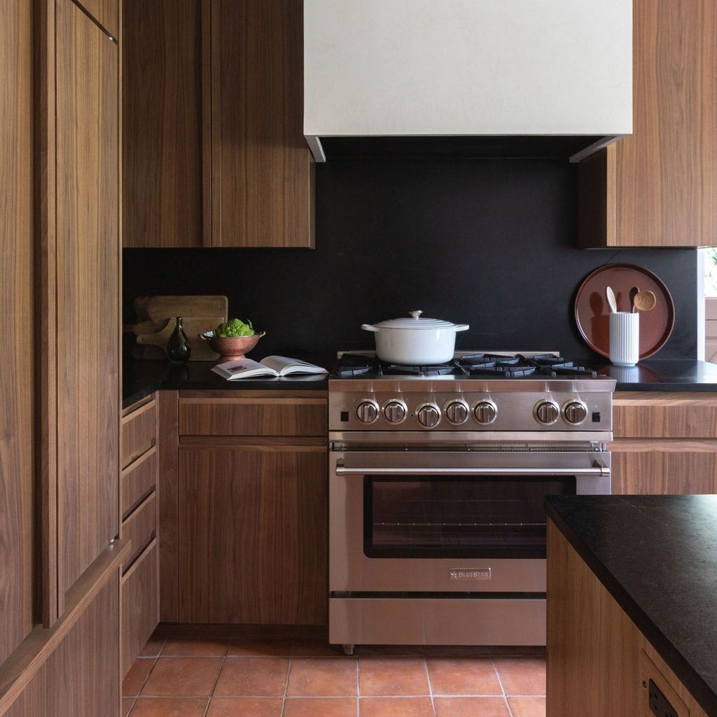 A modern kitchen with wood cabinets, a stainless steel stove with a white pot on top, black countertops, and terracotta tile flooring. A bowl of vegetables, cookbook, and utensils are on the counter.