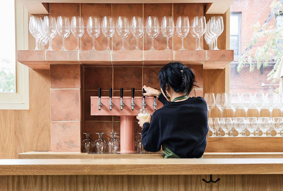 A person with dark hair pours a drink from a row of taps behind a bar, surrounded by neatly arranged wine glasses on shelves. The bar features light wood and brick accents.