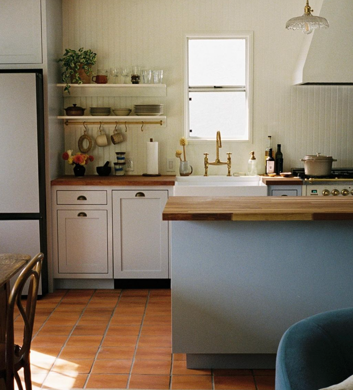 A cozy kitchen with white cabinets, wooden countertops, a farmhouse sink, open shelves with dishes, a window, a stove with a pot, and terracotta tile flooring. A blue chair is partially visible in the foreground.