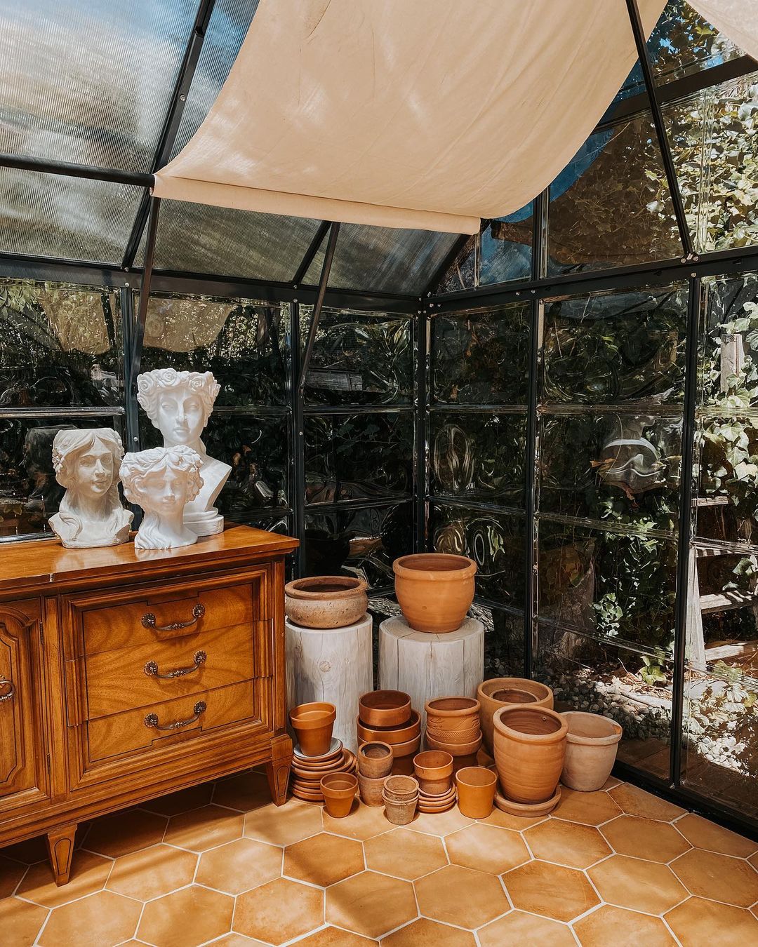 A sunlit greenhouse corner with terra cotta pots of various sizes stacked beside a vintage wooden cabinet topped with two white classical bust sculptures. Greenery is visible through the glass walls.