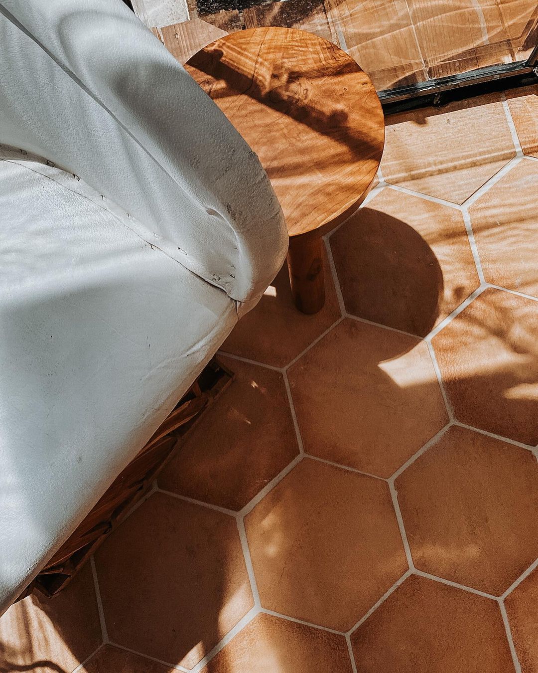 A hexagonal terracotta tile floor with sunlight and shadows. A small round wooden table and a white-cushioned chair are partially visible, creating a cozy and warm atmosphere.