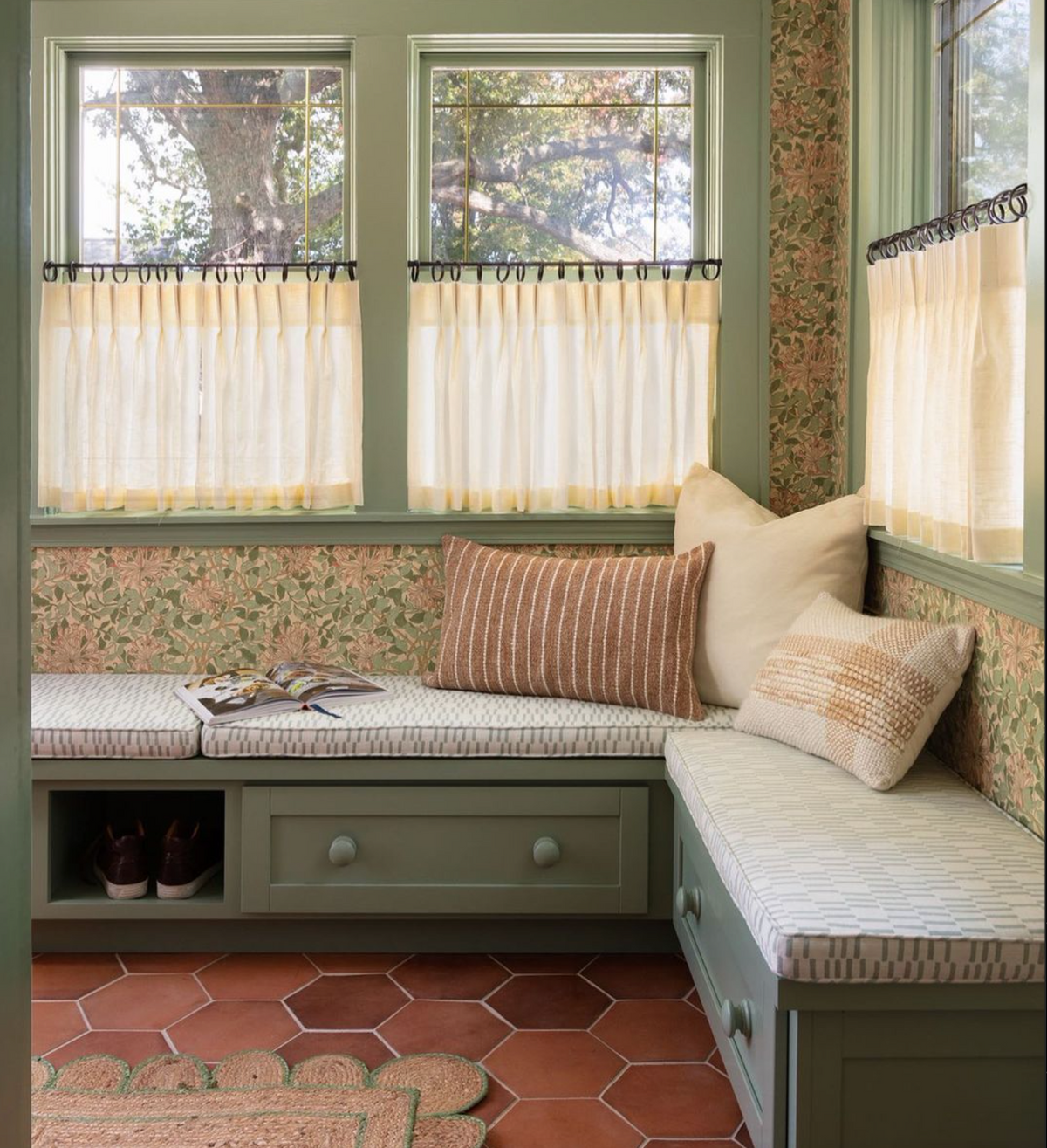 A cozy nook with green built-in benches, patterned cushions, pillows, and storage drawers, near windows with cream curtains. The walls are floral wallpaper, and the floor is terracotta tile. Shoes and a magazine rest on the benches.