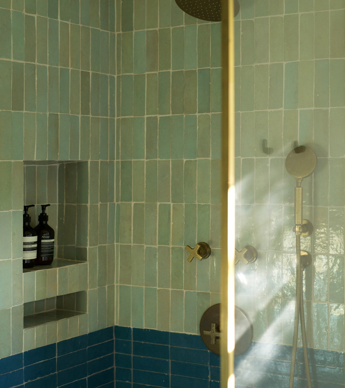 A shower featuring Zia Tile’s Absinthe 2x6 tiles in light green and dark blue, a built-in shelf with two bottles, and brass fixtures—including rainfall showerhead and handheld wand—bathed in sunlight.