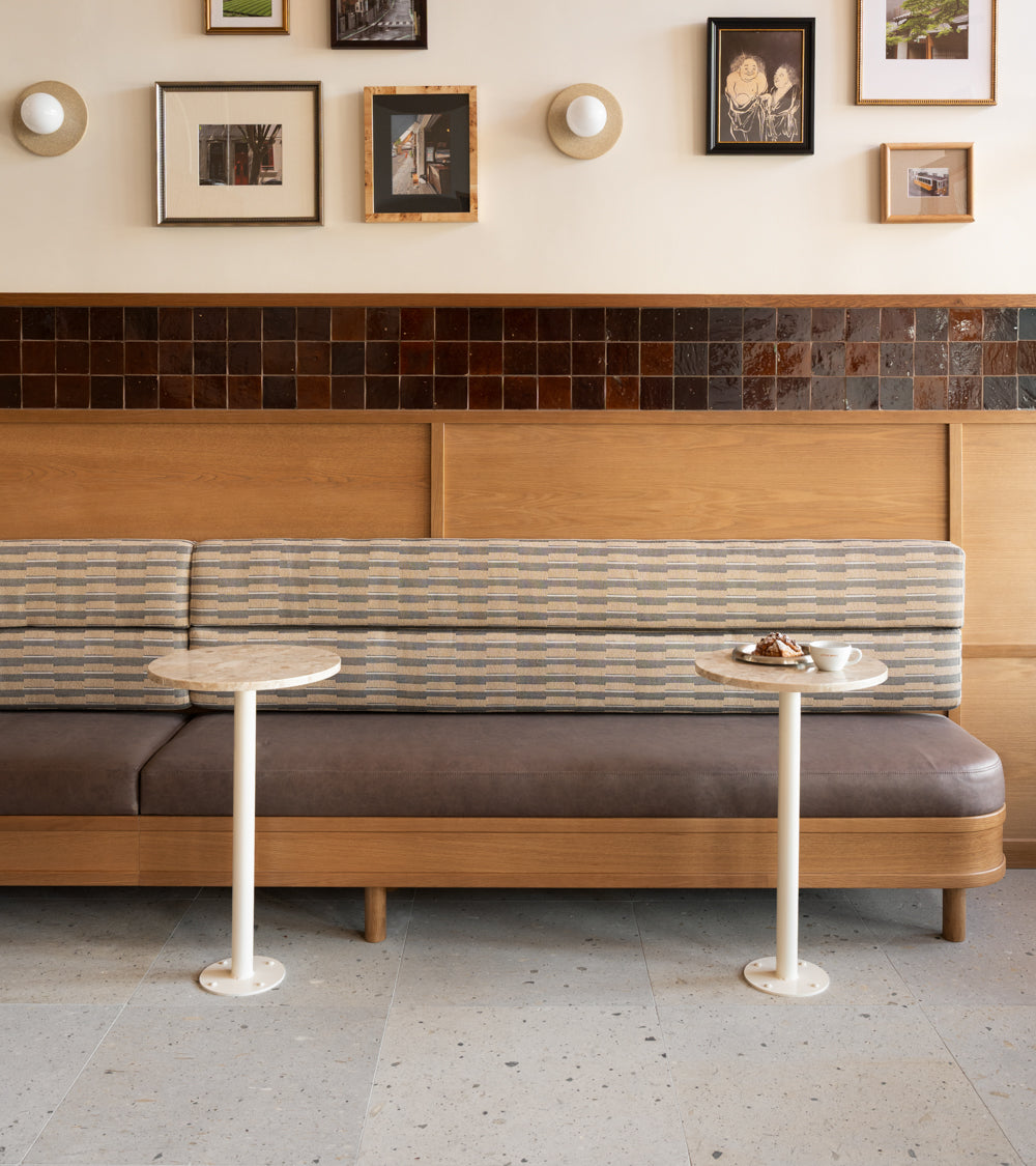 A modern café interior with a long bench, patterned and brown cushions, two small round white tables, Acacia 24x24 tiles from Zia Tile as wall paneling, and framed art on the cream-colored wall above.
