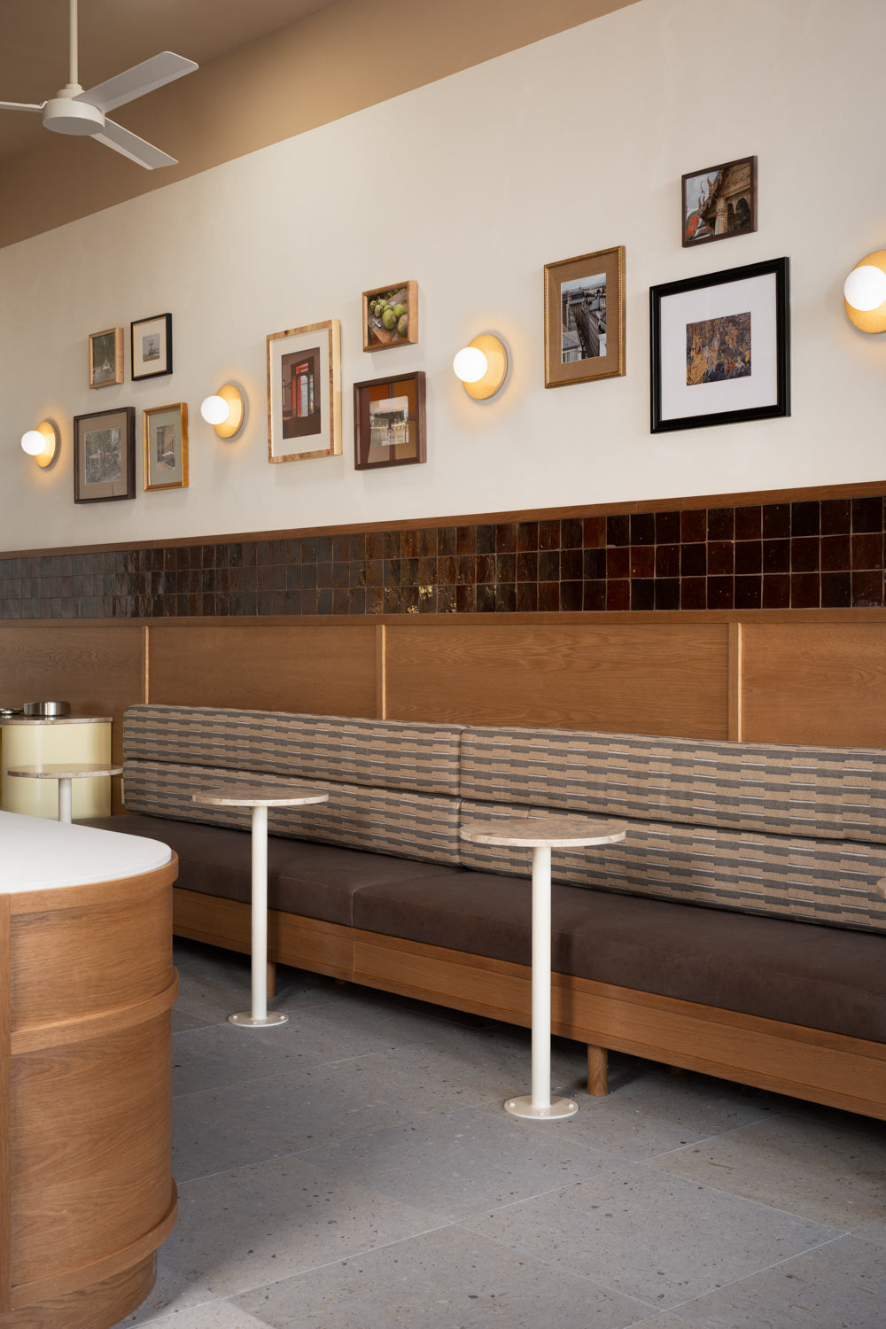 A cozy cafe with a long upholstered bench, small round tables, and framed art above brown and beige Acacia 24x24 tiles by Zia Tile. Wall-mounted lights, a ceiling fan, and part of a counter complete the inviting scene.