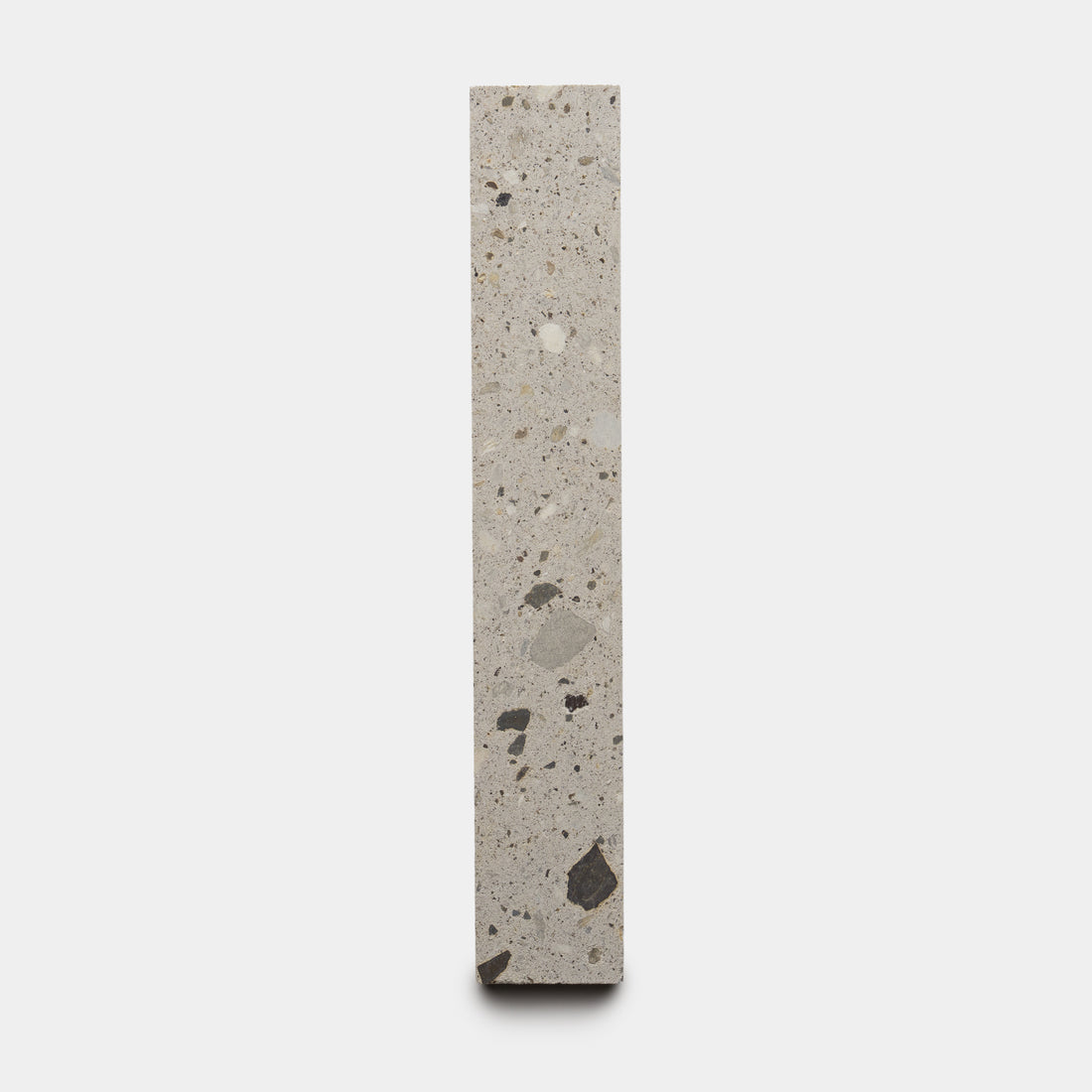The Acacia 2x12 by Zia Tile is a tall, narrow terrazzo tile featuring a light grey background with scattered dark and light stone fragments, shown upright against a plain white background.