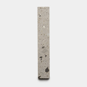 The Acacia 2x12 by Zia Tile is a tall, narrow terrazzo tile featuring a light grey background with scattered dark and light stone fragments, shown upright against a plain white background.