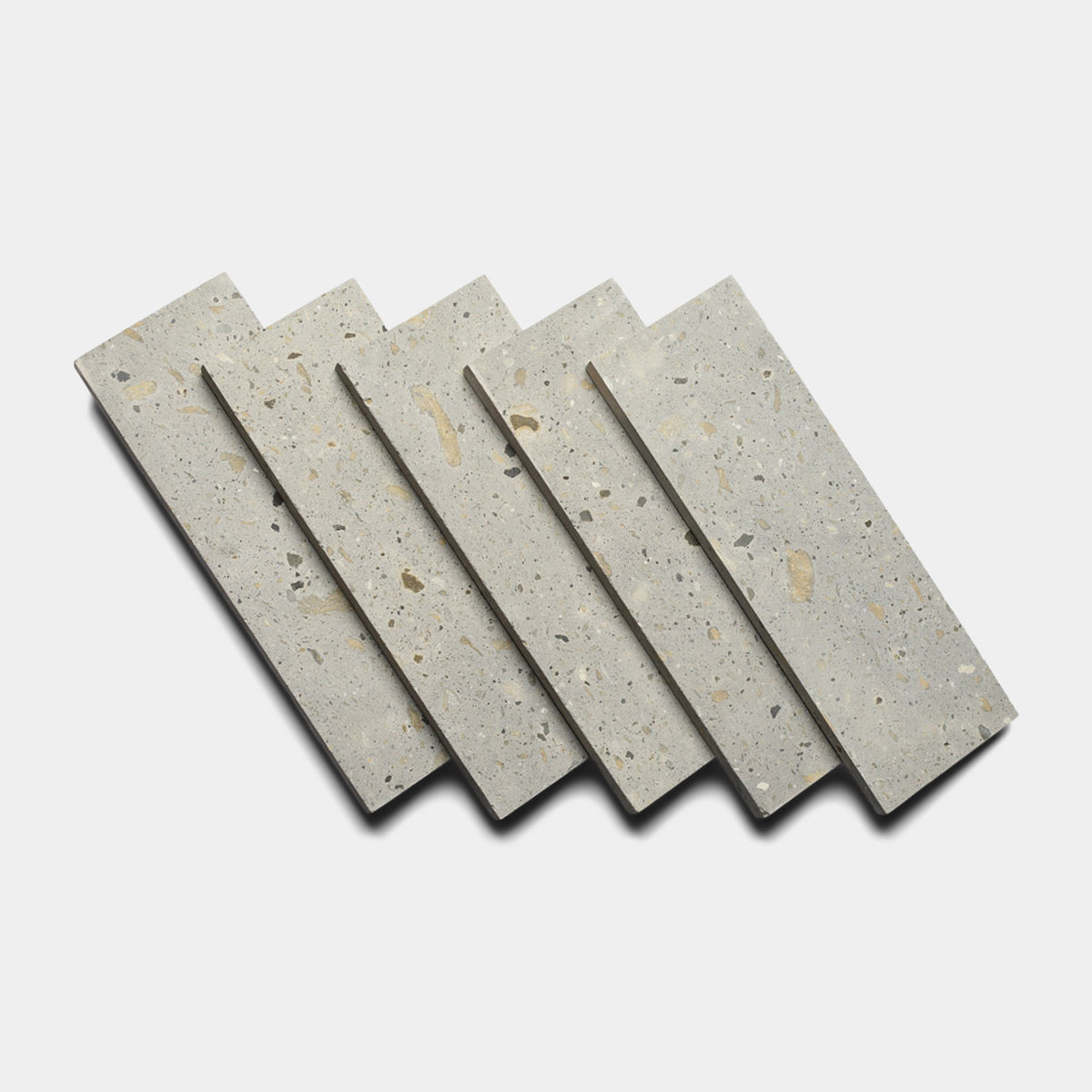 Five Acacia 4x12 rectangular terrazzo tiles by Zia Tile, in light grey with earth-toned aggregates, are arranged in a slightly overlapping diagonal pattern on a white background.