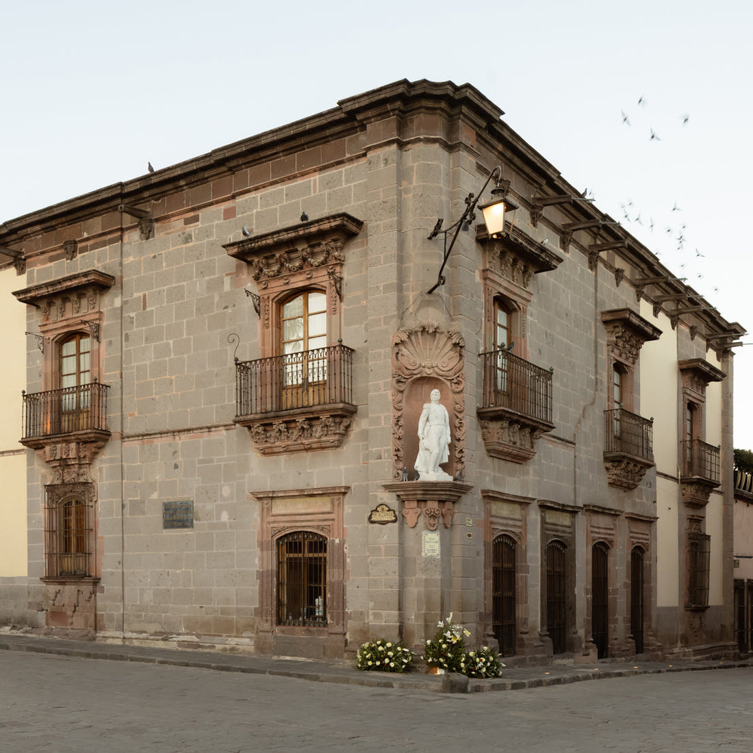 A historic stone building with ornate balconies and large windows, featuring a white statue in an arched niche on the corner. The street is empty, and birds are flying in the sky.