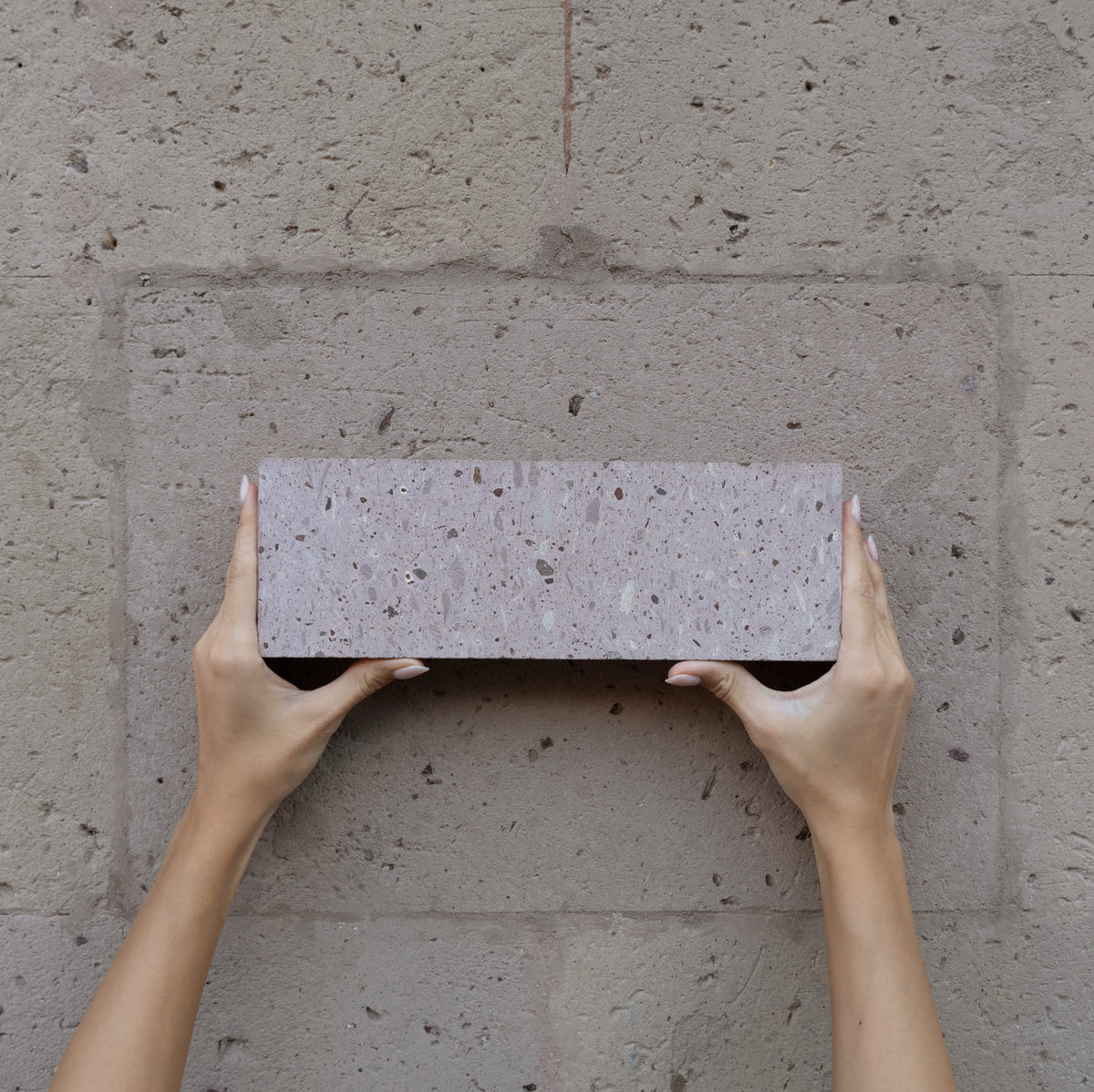 Two hands with manicured nails hold up a rectangular brick against a textured stone wall that matches the color of the brick.