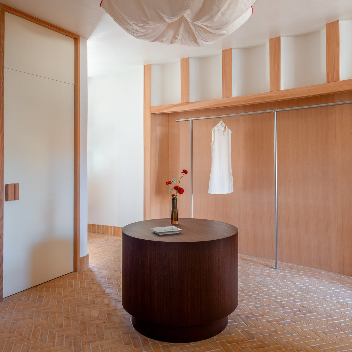A minimalist room with light wood walls and herringbone floor features a round dark table with flowers in a handmade Cotto vase and a book. In the background, a white dress hangs by 2x6 Rectangle + Adobe tiles from Zia Tile.