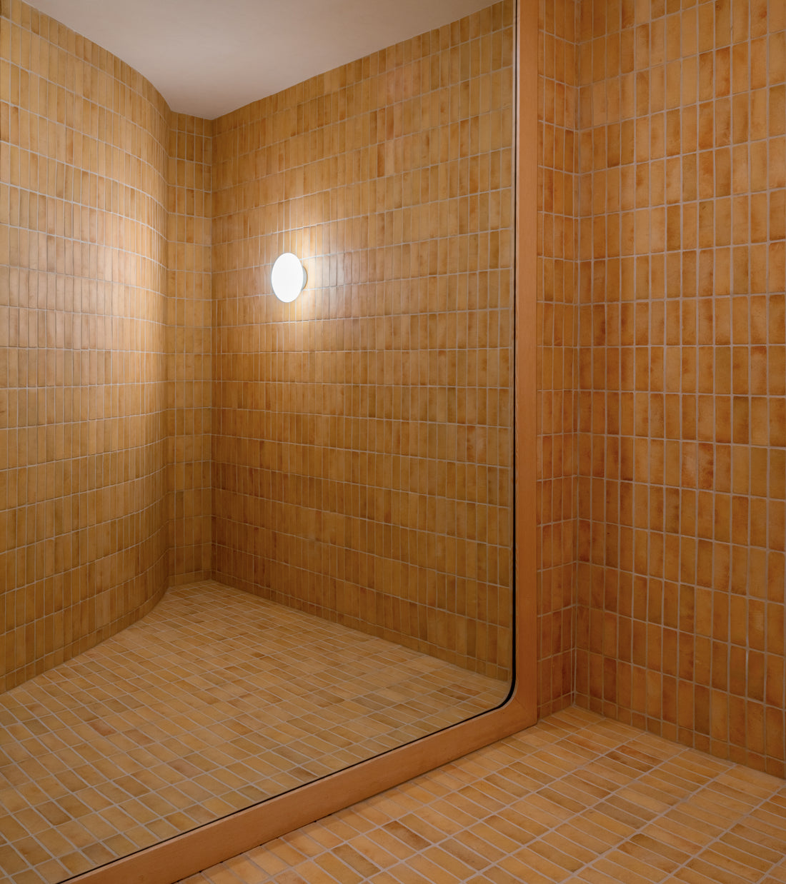 A minimalist room with warm Adobe 2x6 Rectangle tiles by Zia Tile on the floor and walls, featuring a large mirror and a round wall light reflected in the mirror.