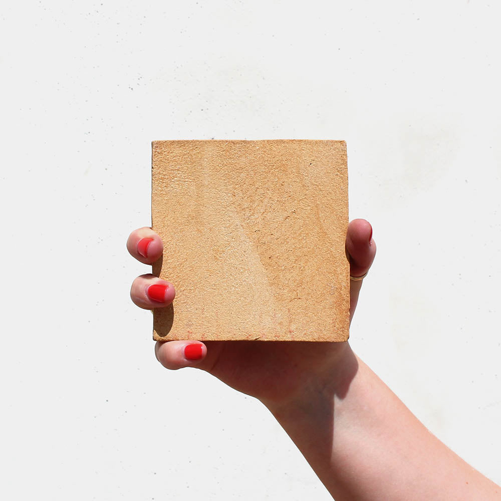A hand with red-painted nails holds up Zia Tile's 4x4 Square + Adobe tile, its light brown color standing out against a plain white background.