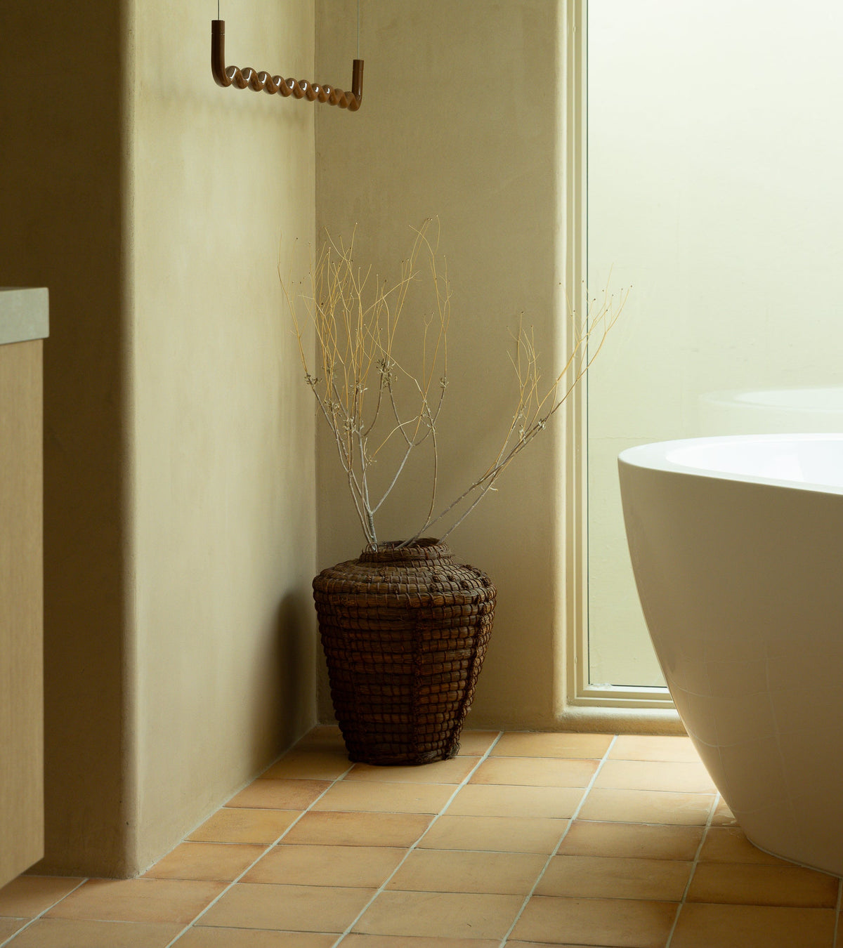 A large woven vase with dry branches stands on a tiled floor next to a modern white bathtub and a tall window in a minimalist, beige-toned bathroom.