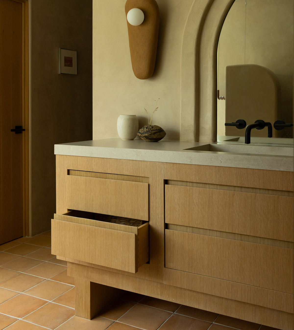 A modern bathroom with a wooden vanity, open drawer, minimalist decor, and an arched mirror features Zia Tile's 8x8 Square + Adobe tiles. Handmade cotto accents inspired by Mexican ceramic arts add warm contemporary style.