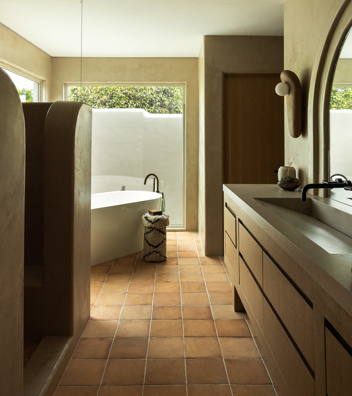 A modern bathroom with terracotta floor tiles, a freestanding white bathtub near large windows, a double sink with wood cabinetry, and curved walls creating a shower area. Natural light fills the space.